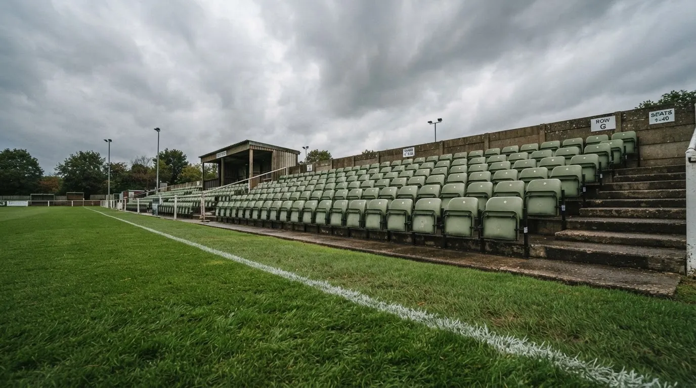Rzędy pustych plastikowych krzesełek małego stadionu widziane z poziomu murawy pod pochmurnym niebem.
