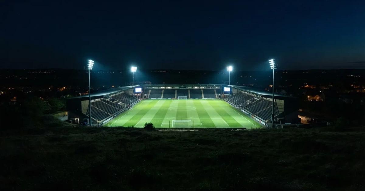 Stadion piłkarski nocą widziany z dużej odległości, jasne reflektory nad murawą i ciemne niebo wokół.