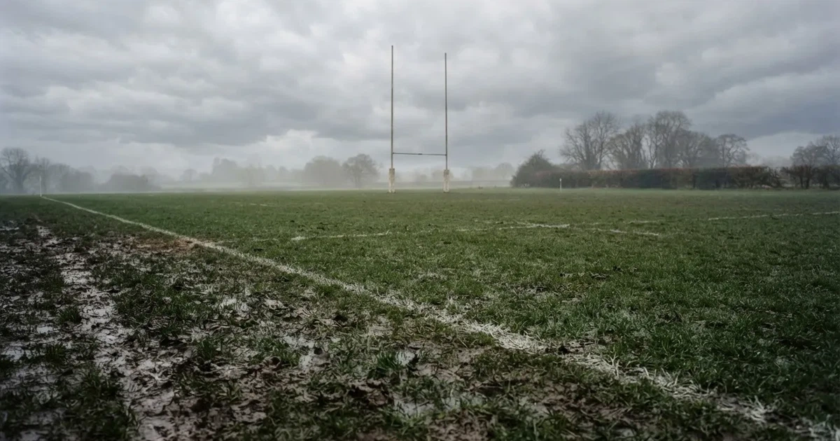 Boisko do rugby ze słupami bramki w oddali pod pochmurnym niebem, mokra trawa i ślady butów.