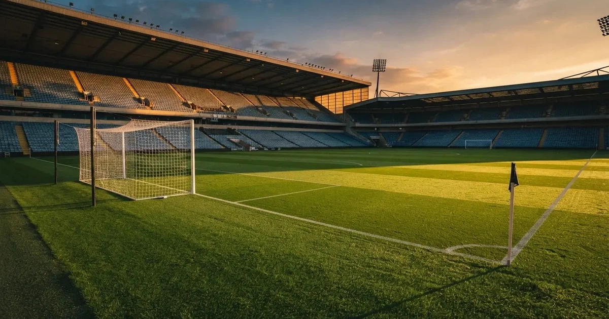 Murawa stadionu o zachodzie słońca z długimi cieniami słupków bramki, trybuny w ciepłym świetle.