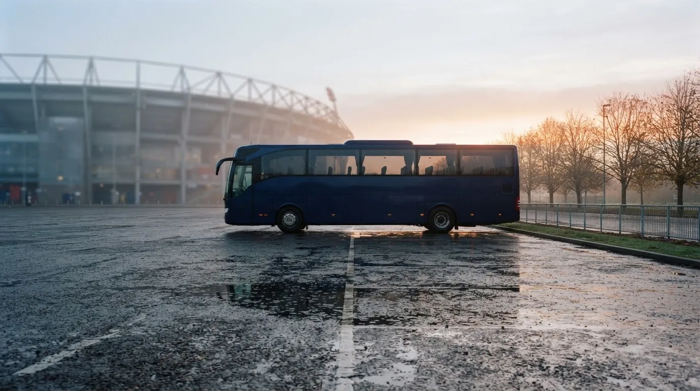 Autokar stoi na parkingu przed stadionem o świcie, rozmyte kontury trybun w tle, pusty plac i mokry asfalt.