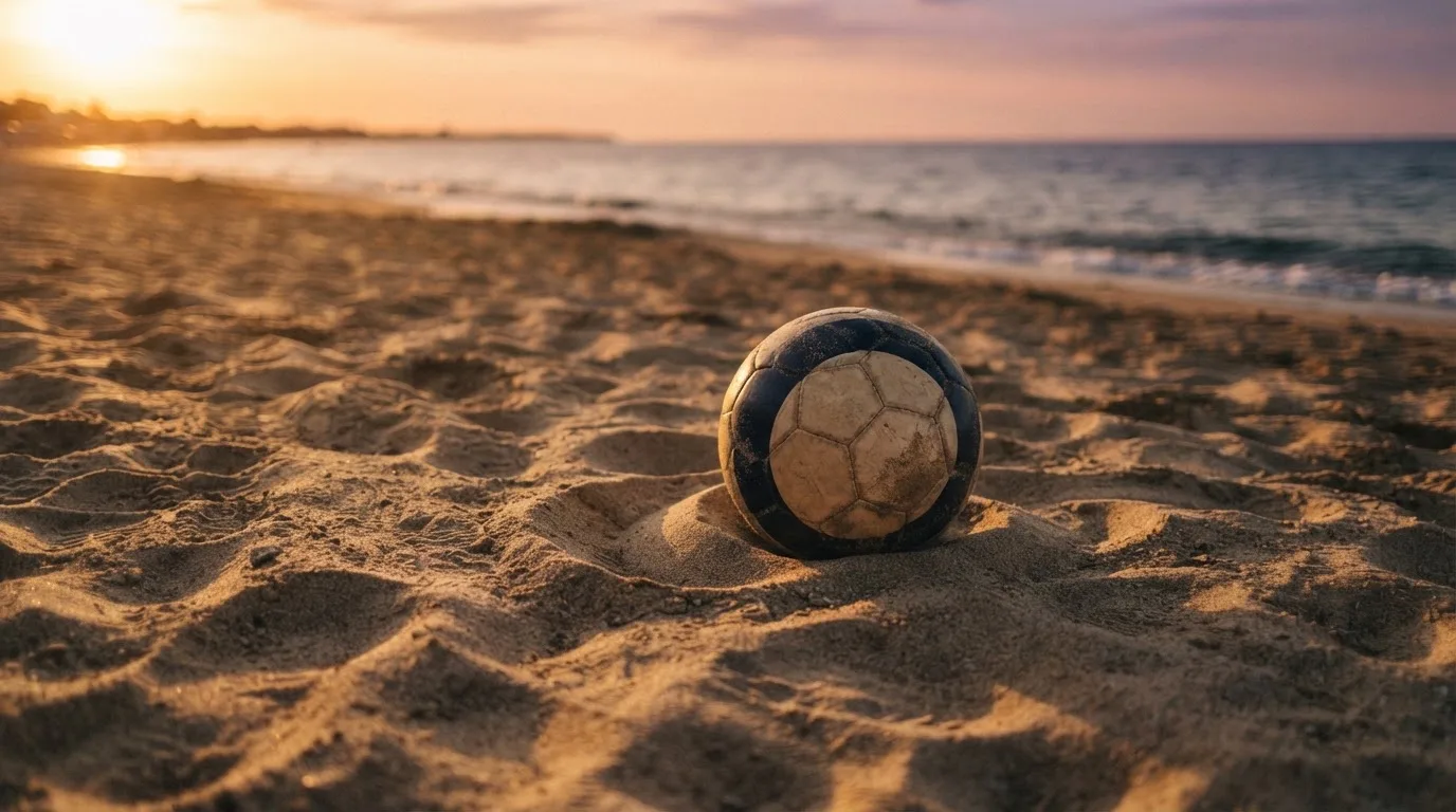 Piłka futbolowa leży na piasku boiska plażowego w ciepłym świetle zachodu, w tle morze i niski horyzont.
