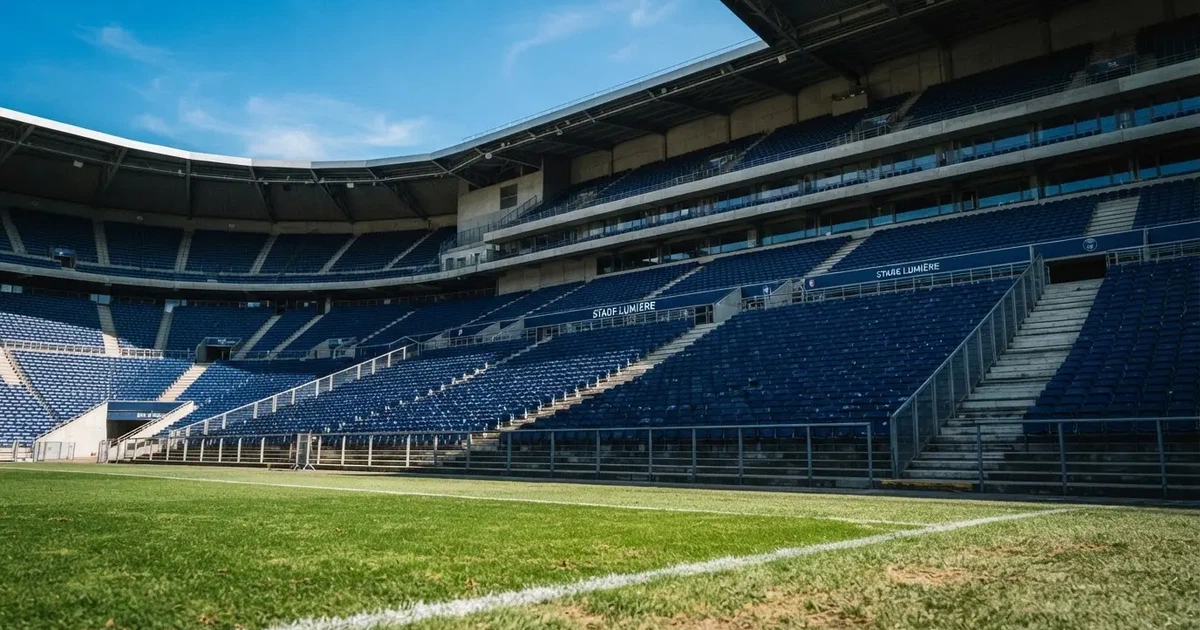 Trybuny wielkiego stadionu widziane z murawy, rzędy pustych ciemnoniebieskich krzesełek, ostre światło dnia i długie cienie.